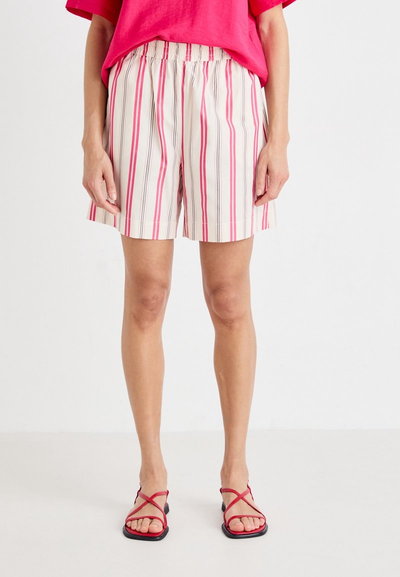 Shorts légers de couleur crème avec des rayures verticales roses et grises. Dotés d'une ceinture élastique et d'une coupe décontractée. Portés avec des sandales rouges à bretelles croisées.