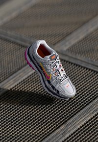 Zapatilla de running Nike con parte superior de malla gris, acentos en púrpura y amarillo, detalles reflectantes y una suela acolchada. Los cordones son grises con toques de naranja.