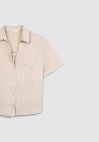 Camicia a maniche corte beige con bottoni in tessuto testurizzato, colletto a punte e due taschini sul petto. Vestibilità rilassata, design accorciato.