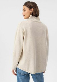 Beige gestrickter Pullover mit hohem Kragen, lässiger Passform, gerippten Bündchen und Saum, kombiniert mit blauen Denim-Jeans. Einfaches und strukturiertes Design.