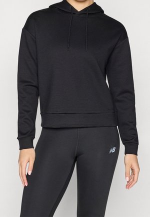 Kvinde iført en sort hættetrøje og sorte træningsleggings med et lille New Balance-logo på låret, stående mod en ensfarvet baggrund.