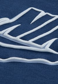 Μπλε βαμβακερό ύφασμα με λευκό ανάγλυφο λογότυπο με ανυψωμένο περίγραμμα, που παρουσιάζει ένα ομαλό και περίπλοκο σχέδιο.