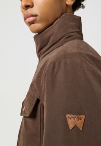 Giacca di stoffa marrone con colletto, dotata di tasca sul petto sinistro e un'etichetta in pelle con il logo Wrangler sulla manica. Texture liscia.