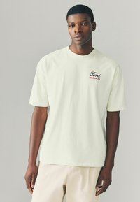 Tricou din bumbac verde deschis, cu mâneci scurte, având un logo "Ford Bronco" negru și roșu pe piept. Modelul stă în picioare, purtând pantaloni bej.