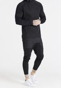 Svart idrottsjacka med dragkedja, strukturerad tyg och logotyp på bröstet, ihop med svarta smala joggingsbyxor och vita sneakers.