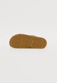 Semelle en caoutchouc brun avec un motif en zigzag, comportant le nom de la marque "TOMS" en relief. Surface lisse avec une texture durable.