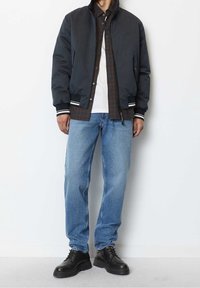 Giacca bomber nera con polsini a righe, camicia a quadri indossata sotto, t-shirt bianca, jeans azzurri e scarpe nere con lacci.