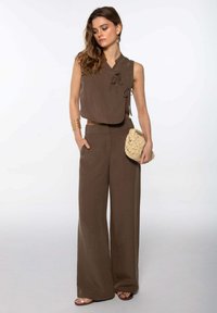 Top corto marrón con detalles de lazo combinado con pantalones anchos marrones. El conjunto presenta una textura natural y se complementa con un bolso de mano tejido en beige.