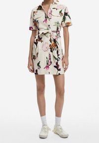 Sukienka w kwiatowy wzór w kolorze kremowym, z krótkimi rękawami, zapinana na guziki z przodu, z paskiem w talii i dwiema przednimi kieszeniami. Uzupełniona o białe skarpetki i jasne buty.