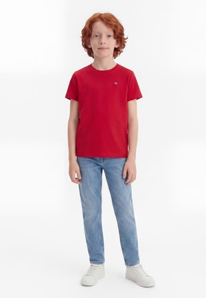 Kind mit roten Haaren, das ein rotes T-Shirt, hellblaue Jeans und weiße Turnschuhe trägt, vor einem schlichten weißen Hintergrund stehend.