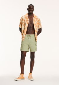 Korte mouw knopsluiting overhemd in crème met een oranje bloemenpatroon, gecombineerd met olijfgroene cargo shorts en oranje hoge sneakers.