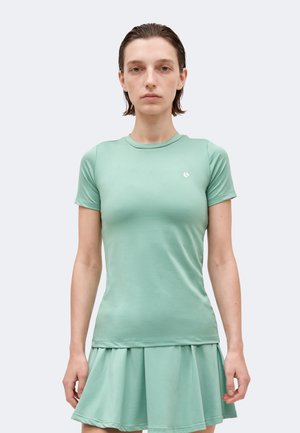 Γυναίκα με κοντά καστανά μαλλιά φοράει ένα εφαρμοστό τοπ μέντας με κοντά μανίκια και ταιριαστό πλισέ φούστα, όρθια μπροστά από ένα απλό φόντο.