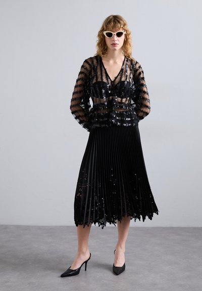 Fekete, áttetsző felső vízszintes flitterekkel, párosítva egy pliszírozott fekete szoknyával, amely csipkés szegéllyel rendelkezik. Fekete tűsarkú cipővel és macska-szem napszemüveggel viselve.