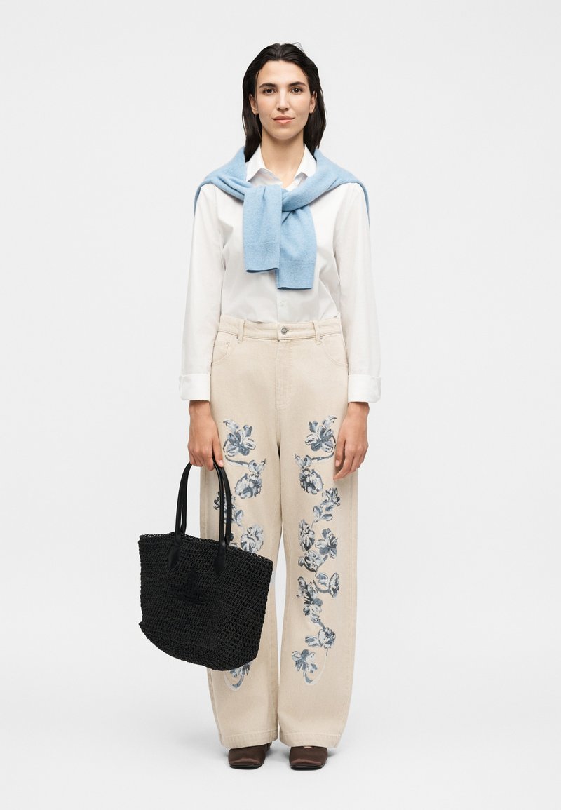 Kvinde iført en hvid skjorte, beige bukser med blå blomsterprint, blå sweater hængt over skuldrene, der holder en sort vævet tote taske.
