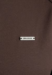 Camiseta de tela marrón con una textura suave, que presenta una etiqueta metálica en tono plateado grabada con "SMILODOX" unida en la parte delantera.