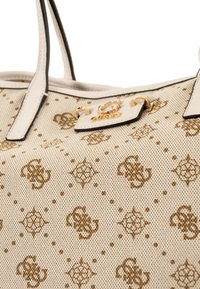 Borsa tote beige con un tessuto testurizzato che presenta un motivo e un logo marroni. Include manici bianchi e un dettaglio in metallo dorato.