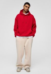 Rode oversized hoodie met een voorzak, trekkoord en ribgebreide manchetten, gecombineerd met losse beige broek en grijze sneakers. Eenvoudig en casual ontwerp.