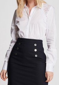White blouse with lace shoulders, button-up front, paired with a high-waisted navy skirt featuring shiny silver buttons.