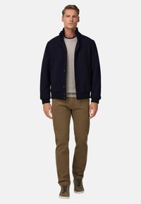 Navy modrá vlna smíšená bunda s vysokým límcem, zapínáním na knoflíky a žebrovanými manžetami, v kombinaci se světle šedým svetrem a hnědými kalhotami. Šedé tenisky.