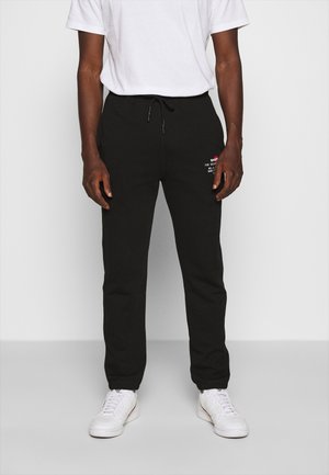 Uomo che indossa pantaloni jogger neri con vita con coulisse e piccolo logo bianco e rosso sulla coscia, abbinati a scarpe da ginnastica bianche e t-shirt bianca.