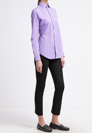 Kobieta ubrana w dopasowaną lawendową koszulę zapinaną na guziki, czarne krótkie spodnie i czarne mokasyny, stojąca na białym tle.