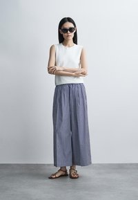 Femme portant des lunettes de soleil noires, un haut blanc sans manches, un pantalon large à rayures verticales bleu et blanc, et des sandales marron, debout les bras croisés.