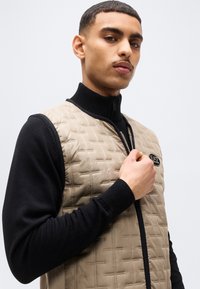 Άνδρας που φοράει μπεζ γιλέκο με καπιτονέ σχέδιο πάνω από μαύρο μπλουζάκι με μακριά μανίκια, κρατώντας το φερμουάρ κοντά στο στήθος, κοιτάζει προς την κάμερα με ουδέτερη έκφραση.