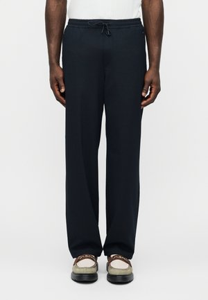 Mees, kes kannab lahtisi musta värvi pükse, millel on nöörvöökoht, ning beeže loafer-kingi pruunide rihmadega, seisab valge tausta ees.