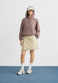 Νεαρή γυναίκα που φοράει μια μωβ φούτερ με κουκούλα, μπεζ φούστα, λευκές κάλτσες, μπεζ αθλητικά παπούτσια και μπεζ καπέλο μπέιζμπολ της LA σε μπλε δάπεδο.