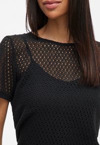 Svart strikket topp med et åpent, teksturert mønster, rund halslinning og korte ermer. Stoffet er lett og pustende.