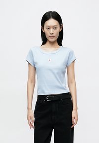 Light blue ribbed t-shirt with short sleeves, rounded neck, and a small red and white logo in the center, paired with black high-waisted pants.