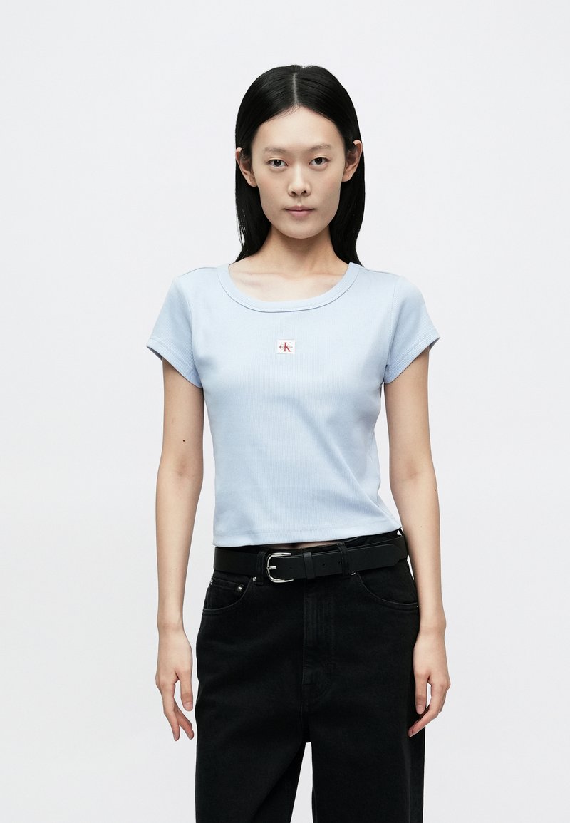 Light blue ribbed t-shirt with short sleeves, rounded neck, and a small red and white logo in the center, paired with black high-waisted pants.