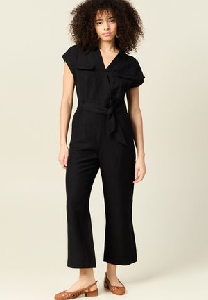 Mono de lino negro con mangas cortas, diseño envolvente, lazo en el frente y pantalones de pierna ancha. Se lleva con zapatos de lona trenzados de color marrón claro.