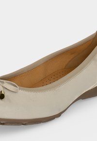 Chaussure plate beige avec un petit nœud et un accent doré sur l'avant, semelle intérieure perforée et rembourrée, semelle en caoutchouc marron.