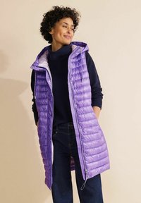 Lila pufferväst med vertikal sömnad, dragkedja fram och justerbar huva, bärs över en mörk tröja och jeans mot en ljus bakgrund.