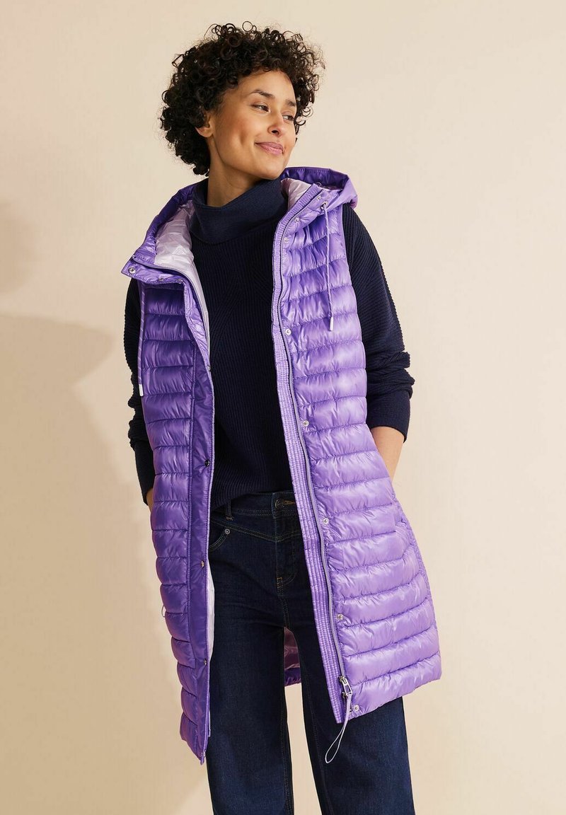 Lila pufferväst med vertikal sömnad, dragkedja fram och justerbar huva, bärs över en mörk tröja och jeans mot en ljus bakgrund.