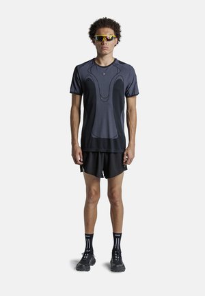 Uomo che indossa una maglietta sportiva nera, pantaloncini, scarpe da corsa nere, calzini neri e occhiali da sole sportivi con lenti gialle, in piedi su sfondo bianco.