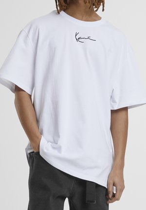 Personne portant un t-shirt blanc ample avec un petit logo signature noir près de l'encolure, assorti à un pantalon foncé, une main dans la poche.