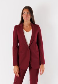 Donna in piedi con un completo bordeaux, giacca a un bottone e top bianco, che guarda leggermente di lato su uno sfondo bianco semplice.