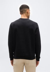 Άνδρας που φοράει μαύρο μακρυμάνικο φούτερ και μπεζ παντελόνι, γυρισμένος με την πλάτη προς ένα απλό, φωτεινό φόντο.