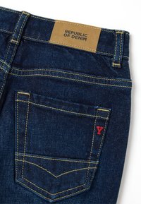 Jeans de mezclilla azul oscuro con una etiqueta color beige que dice "REPÚBLICA DE MEZCLILLA", costuras amarillas y un acento rojo en forma de "Y" en el bolsillo trasero.