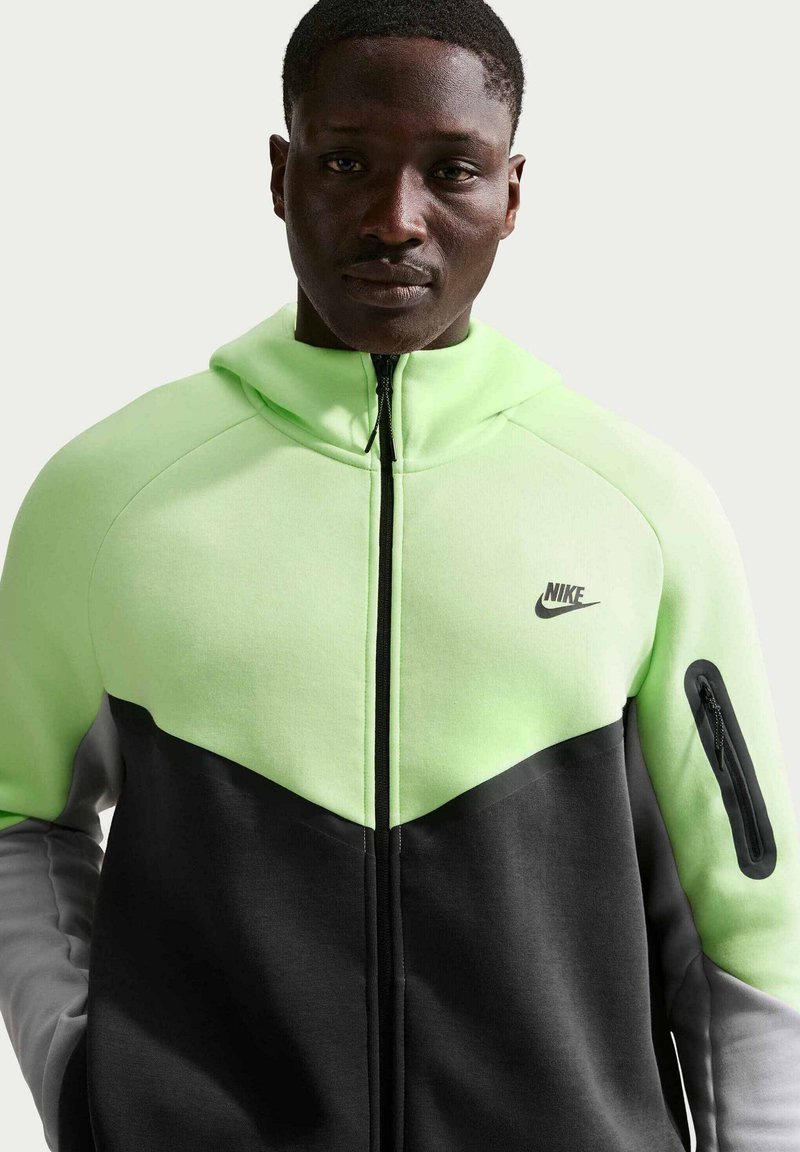 Mand iført en neongrøn og sort Nike lynlåssweatshirt med lynlåslomme på venstre ærme, set forfra mod en ensfarvet baggrund.