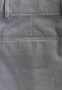 Tela de pana gris con costillas verticales, que presenta un bolsillo trasero con un contorno cosido. Textura suave y color uniforme.