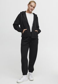 Hamac à capuche noir avec poches et cordons blancs, associé à un pantalon de survêtement noir et des baskets blanches. Design simple et décontracté avec une coupe relaxée.