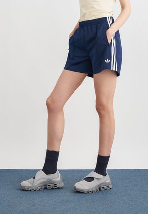 Person iført marineblå Adidas-shorts med hvide striber, sorte sokker og grå sportssko, der står på blå tæppe.