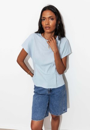 Femme aux longs cheveux foncés portant une chemise bleu clair à manches courtes et un short en jean bleu jusqu’aux genoux, debout contre un mur blanc.