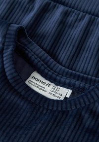 Tuvo plāna skata uz ribota navy blue auduma apkakles, ar iešūtu balto apģērba etiķeti, kurā norādītas izmēra un izcelsmes detaļas.