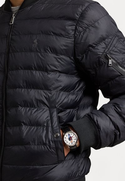 Svart boblejakke med horisontale sømmer og ribbestrikkede mansjetter, med en glidelåslomme. Et rundt klokke med hvit urskive og svart rem er synlig.