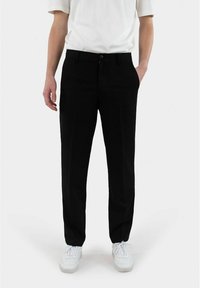 Schwarze maßgeschneiderte Hose mit geradem Bein, Fronttaschen und Knopfverschluss, kombiniert mit einem weißen kurzärmeligen Hemd und Turnschuhen.
