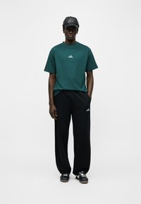 Άνδρας στέκεται με τα χέρια στις τσέπες, φορώντας μπλούζα σκούρου πράσινου χρώματος, μαύρο φόρμα παντελόνι, μαύρα αθλητικά παπούτσια και μαύρο δερμάτινο καπέλο μπέιζμπολ, μπροστά σε λευκό φόντο.